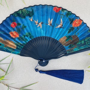 Puede incluir: Un abanico azul y verde con un mango negro y una borla azul. El abanico presenta un dise&ntilde;o floral con flores rojas y p&aacute;jaros volando.