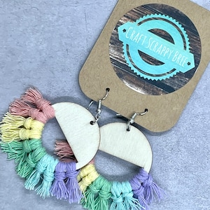 Puede incluir: Pendientes de borla de hilo de colores arcoíris con bases de madera semicirculares. Los pendientes están unidos a ganchos plateados.