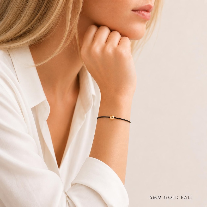 Minimalist black cord bracelet with 5mm gold ball worn by a blonde woman in a white blouse, adjustable waterproof bracelet handmade by Lucky Charms USA