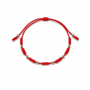 May include: A red string bracelet featuring a series of small, polished gold-tone spherical beads interspersed with knotted sections of the red cord. The bracelet includes an adjustable sliding knot closure with two dangling ends tipped with gold beads.