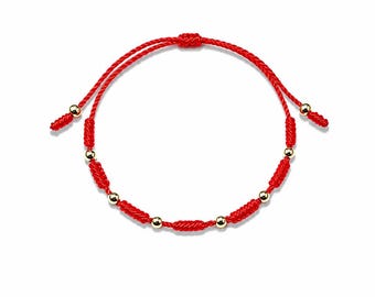 May include: A red string bracelet featuring a series of small, polished gold-tone spherical beads interspersed with knotted sections of the red cord. The bracelet includes an adjustable sliding knot closure with two dangling ends tipped with gold beads.
