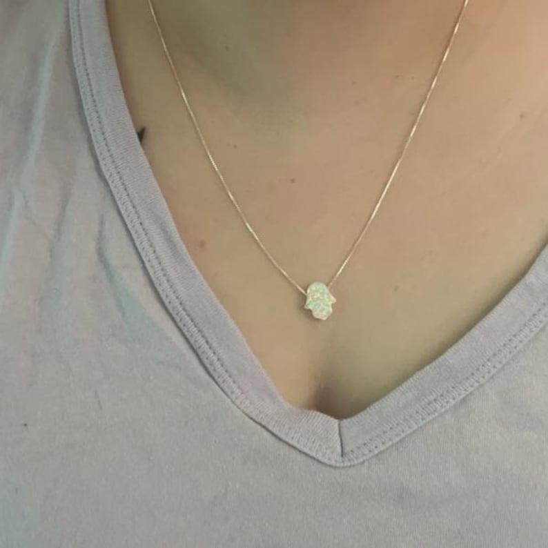 Pode incluir: Um delicado colar de corrente dourada com um pequeno pingente de m&atilde;o Hamsa branco, semelhante ao opala. O pingente est&aacute; centrado no peito, e o colar &eacute; usado sobre uma camiseta lil&aacute;s clara.