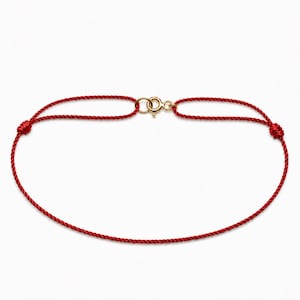 May include: A thin red cord bracelet featuring a gold-toned metal spring ring clasp. The bracelet is constructed from a single continuous strand of red thread that forms a loop, secured at the top by the small circular gold-coloured metal fastener.