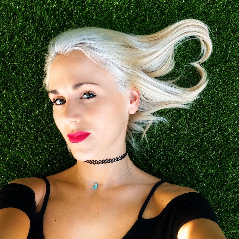 Lifestyle photo of a woman lying on green grass wearing a dainty moon opal necklace on a delicate chain with a black top and soft natural outdoor styling. lucky charms usa
