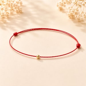 May include: A minimalist red string bracelet with a small gold bead in the center. The bracelet is tied with knots on each end. The background is a soft, neutral color with delicate white flowers.