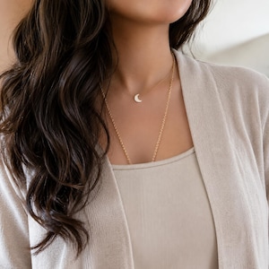 Lifestyle photo of a woman with long wavy brunette hair wearing a dainty white crescent moon necklace by lucky charms usa layered with a delicate chain and styled with modern neutral clothing in soft natural indoor light.