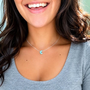 Close-up lifestyle photo of a smiling woman wearing a dainty blue opal crescent moon necklace on a delicate chain by lucky charms usa with soft natural lighting and casual everyday styling.