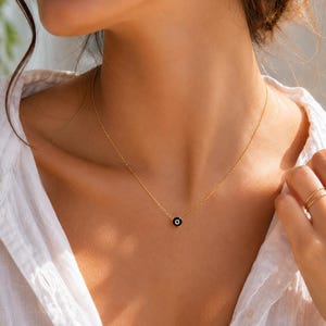 Puede incluir: Un collar de cadena dorada fina con un pequeño colgante circular que presenta un fondo de esmalte negro y un punto blanco central, similar a un motivo de ojo. El collar descansa sobre la clavícula, mostrando un diseño sencillo.