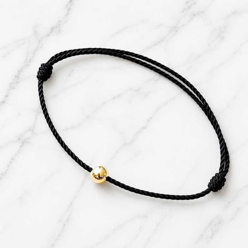 Minimalist black cord bracelet with polished gold ball on white marble background, adjustable waterproof bracelet handmade by Lucky Charms USA