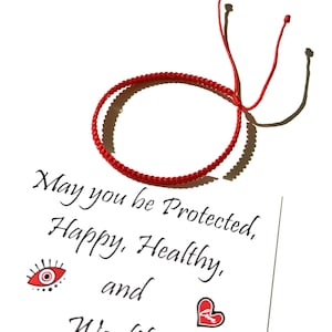 Peut inclure: Un bracelet rouge tressé avec des liens réglables. Le bracelet est sur un fond blanc avec le texte "May you be Protected, Happy, Healthy, and Wealthy" et un œil rouge et un cœur graphique.