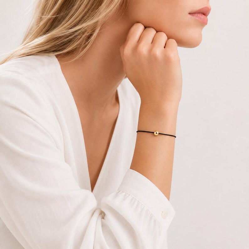 Minimalist black cord bracelet with gold ball worn by a blonde woman in a white blouse, adjustable waterproof bracelet handmade by Lucky Charms USA