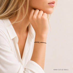Minimalist black cord bracelet with 4mm gold ball worn by a blonde woman in a white blouse, adjustable waterproof bracelet handmade by Lucky Charms USA
