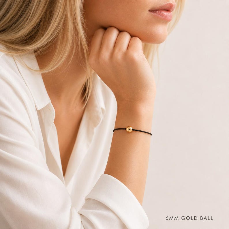 Minimalist black cord bracelet with 6mm gold ball worn by a blonde woman in a white blouse, adjustable waterproof bracelet handmade by Lucky Charms USA