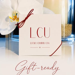 May include: A square white card with red text reading LCU LUCKY CHARMS USA and a small heart symbol. The card is tied with a thin red string and features the red script text Gift-ready for your favorite people with a small heart symbol below.