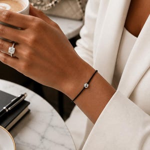 Minimalist silver ball bracelet on black cord worn with a cream blazer at a luxury cafe, adjustable waterproof bracelet handmade by Lucky Charms USA