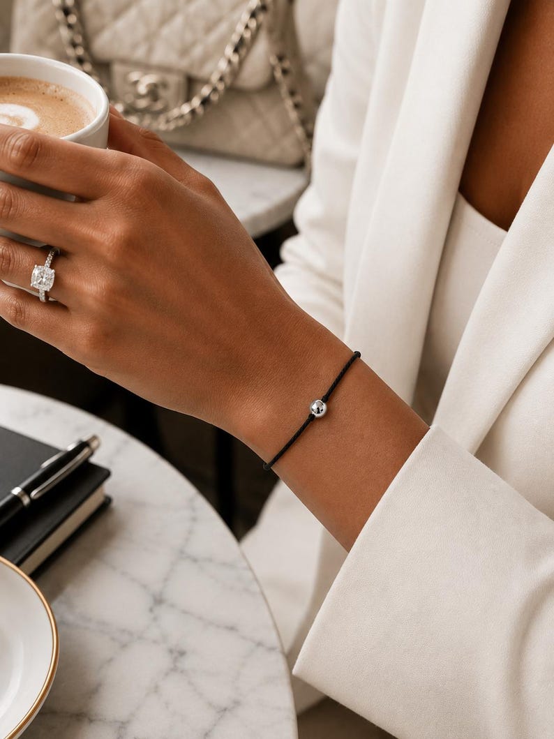 Minimalist silver ball bracelet on black cord worn with a cream blazer at a luxury cafe, adjustable waterproof bracelet handmade by Lucky Charms USA