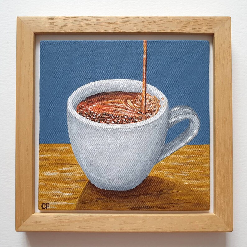 May include: A square painting in a wooden frame depicts a white coffee cup being filled with brown liquid. The background is blue, and the surface beneath the cup is a warm yellow-brown. The artist's initials, "CP," are in the lower left corner.