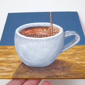 May include: A painting of a white coffee cup with a handle, filled with brown liquid, with a stream of coffee pouring into it. The background is blue and the surface is a light brown color. The painting is on a square canvas.