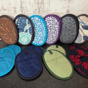 May include: A selection of oval-shaped pot holders in diverse patterns and colours. The pot holders display designs in blue, brown, purple, and green, with floral and abstract motifs. Some have a loop for hanging, suitable for kitchen use.