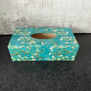 May include: A teal blue tissue box cover with a floral design of white blossoms and branches. The cover has a rectangular opening for tissues.