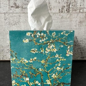 May include: A teal blue tissue box cover with a white floral design. The design features a repeating pattern of white blossoms and branches on a teal blue background.