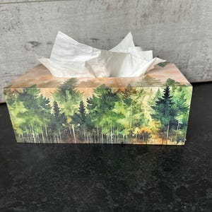 May include: A wooden tissue box cover featuring a watercolour design of a forest scene. The box is decorated with green trees and a light orange background.