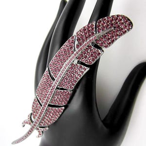 May include: A silver-toned metal brooch shaped like a feather, featuring a detailed quill design. The surface of the feather is densely encrusted with numerous small, round, pink rhinestones that cover the entire front of the piece.