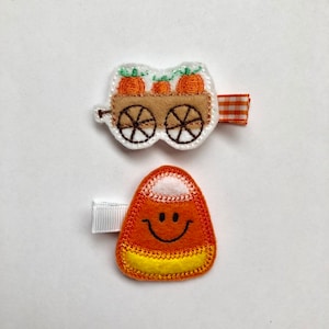 May include: Two felt hair clips with an autumn theme. One clip features a wagon filled with pumpkins and the other clip is a smiling candy corn.