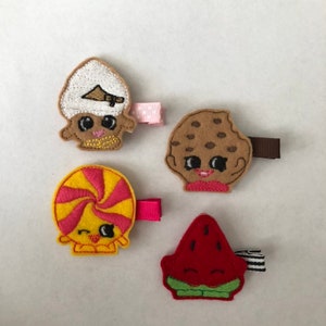 May include: Four felt hair clips with cute cartoon faces. The clips are shaped like a cookie, a watermelon, a candy cane, and a chef's hat.