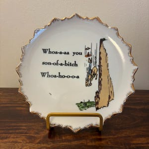 May include: A white decorative plate with a gold rim and a humorous illustration of a cowboy falling off a cliff. The plate has the text "Whoa-a-aa you son-of-a-bitch Whoa-ho-o-o-a" in black font.