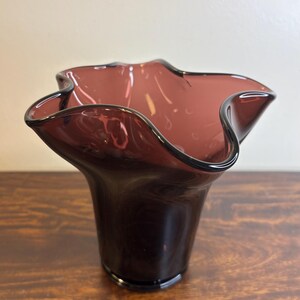 May include: A dark purple glass vase with a ruffled rim. The vase has a flared shape and sits on a wooden surface. The glass is translucent, allowing light to pass through.