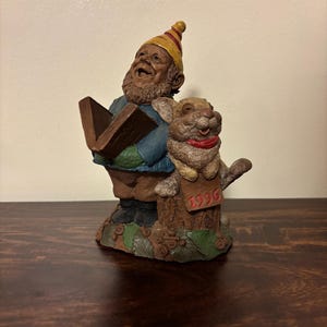 May include: A whimsical figurine of a smiling gnome reading a book, accompanied by a cheerful rabbit. The gnome wears a yellow hat with red stripes, a blue jacket, and black boots. The rabbit has a red collar and a sign that reads "1996".