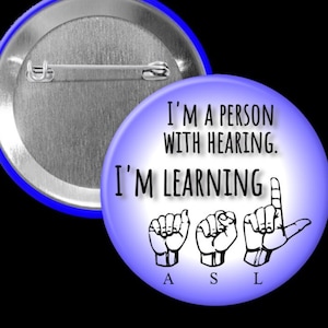 Peut inclure: Un bouton bleu avec un fond blanc et le texte "Je suis une personne avec l'ouïe. J'apprends l'ASL" avec les lettres A, S et L sous le texte. Les lettres sont en langue des signes.