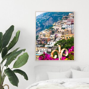 Puede incluir: Una impresi&oacute;n enmarcada de un pueblo costero italiano, con edificios coloridos en una ladera. La imagen muestra una escena vibrante con flores rosas en primer plano y una planta verde en una cesta de mimbre.