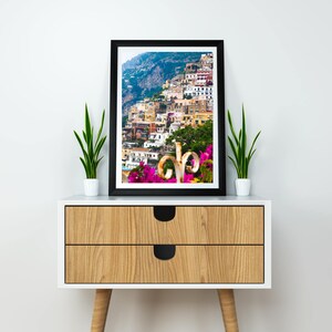 Puede incluir: Una impresi&oacute;n enmarcada de un pueblo costero italiano con edificios coloridos en una ladera. El arte enmarcado en negro se exhibe en un gabinete blanco y de madera, flanqueado por plantas en macetas. El gabinete tiene dos cajones con manijas circulares negras.