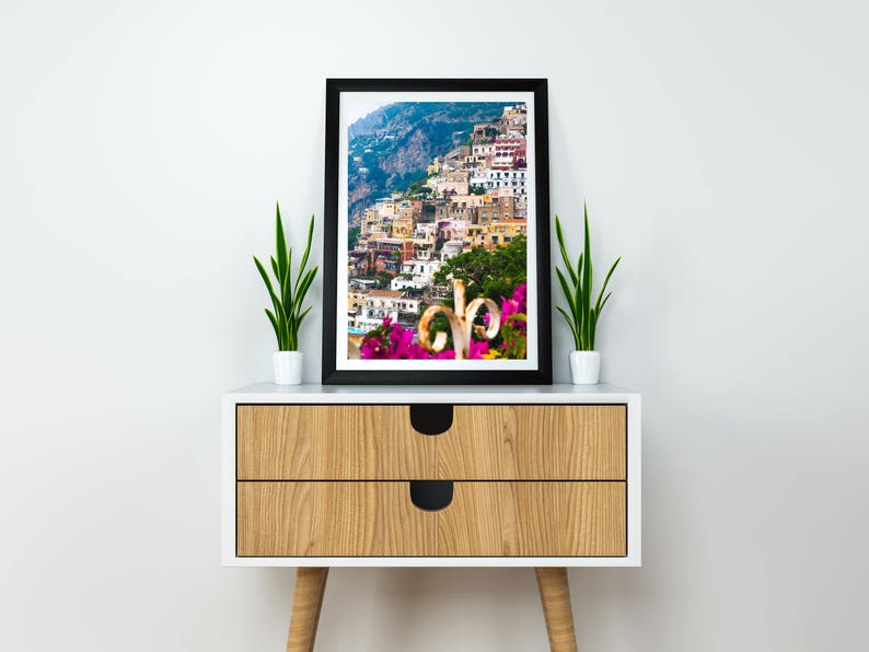 Puede incluir: Una impresi&oacute;n enmarcada de un pueblo costero italiano con edificios coloridos en una ladera. El arte enmarcado en negro se exhibe en un gabinete blanco y de madera, flanqueado por plantas en macetas. El gabinete tiene dos cajones con manijas circulares negras.