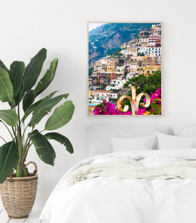 Puede incluir: Una impresi&oacute;n enmarcada de un pueblo costero italiano, con edificios coloridos en una ladera. La imagen muestra una escena vibrante con flores rosas en primer plano y una planta verde en una cesta de mimbre.