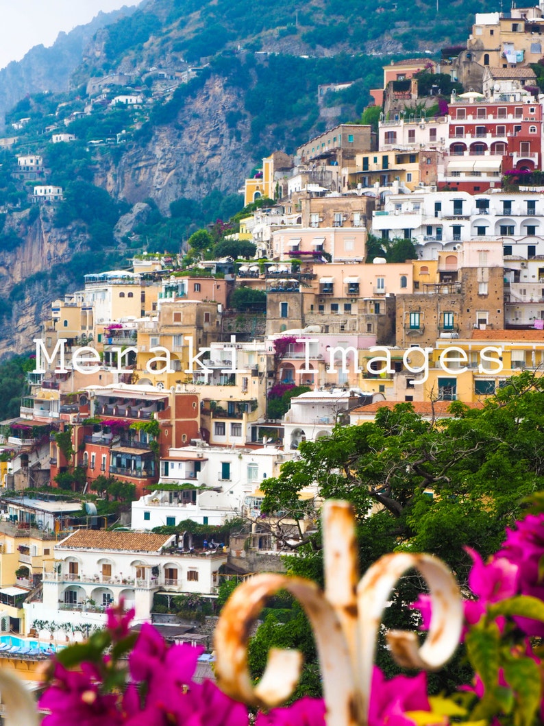 Puede incluir: Una vista pintoresca de un pueblo costero en una ladera. Edificios con fachadas coloridas en tonos amarillos, rosas y blancos descienden en cascada por la ladera. Frondosos &aacute;rboles verdes y vibrantes flores rosas enmarcan el primer plano. Incluye el texto "Meraki Images".