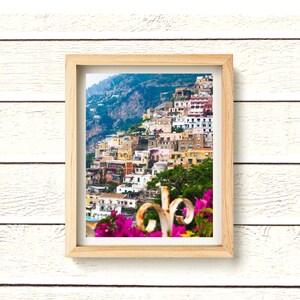 Puede incluir: Una impresi&oacute;n enmarcada de un pueblo costero italiano en una ladera. Los edificios son de colores pastel, con una monta&ntilde;a y un cielo azul como tel&oacute;n de fondo. El marco es de madera clara y el primer plano presenta flores rosas vibrantes.