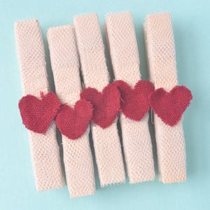 May include: Five wooden clothespins arranged vertically, each wrapped in a textured, light beige fabric ribbon. A small, deep red heart cut from fabric is attached to the center of each clothespin, creating a uniform row of five hearts.