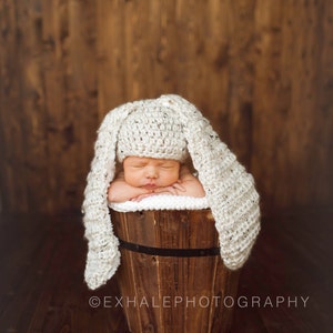 May include: A newborn baby wearing a white crocheted bunny hat sleeps in a wooden bucket. The bucket is on a wooden floor.