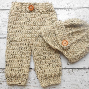 May include: A set of crocheted baby clothes in a beige color with brown flecks. The set includes a pair of pants and a hat. Both pieces have a brown button.