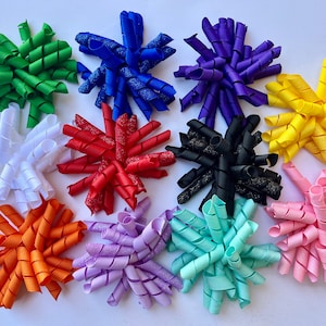 May include: Twelve different colored curly ribbon bows. The bows are made of a variety of materials, including satin, glitter, and velvet. The bows are arranged in a circle on a white background.