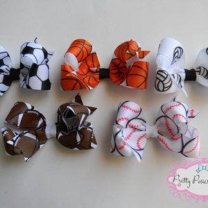 May include: Six hair bows featuring various sports themes. The bows are crafted from white, brown, orange, and red ribbon with black and white printed designs of basketballs, footballs, baseballs, soccer balls, and volleyballs.
