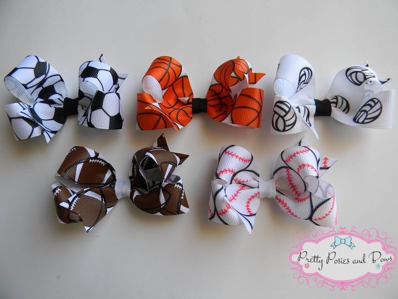 May include: Six hair bows featuring various sports themes. The bows are crafted from white, brown, orange, and red ribbon with black and white printed designs of basketballs, footballs, baseballs, soccer balls, and volleyballs.