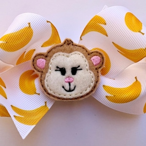 May include: A white hair bow with a pattern of yellow bananas. The bow features a felt monkey face in the center with pink ears and a pink nose. The bow is a fun accessory.