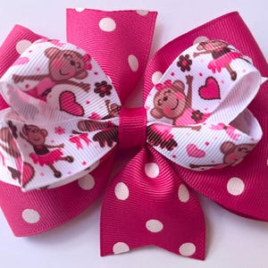 May include: A large, handmade hair bow featuring a layered design. The bow has a vibrant pink base with white polka dots. The center layer is white with a repeating pattern of dancing monkeys, hearts, and flowers. A charming accessory.