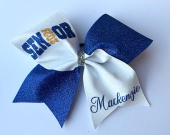 May include: A glittery cheerleading bow in white and blue. The bow features the word "SENIOR 2023" in blue and gold, and the name "Mackenzie" in blue script. A silver rhinestone embellishment is at the center.