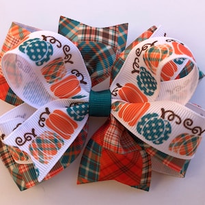 May include: A white, orange, and teal hair bow with a plaid pattern and the text "Happy Fall" printed on it.