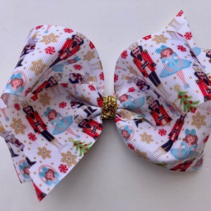 May include: A white, red, and blue bow with a Nutcracker and Sugar Plum Fairy print. The bow has a gold glitter center.